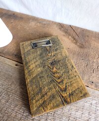 May include: A rectangular wooden clipboard featuring a dark brown stained finish with visible wood grain and a prominent knot. The board is equipped with a metallic bronze-toned spring-loaded clip secured by two small black screws at the top edge.