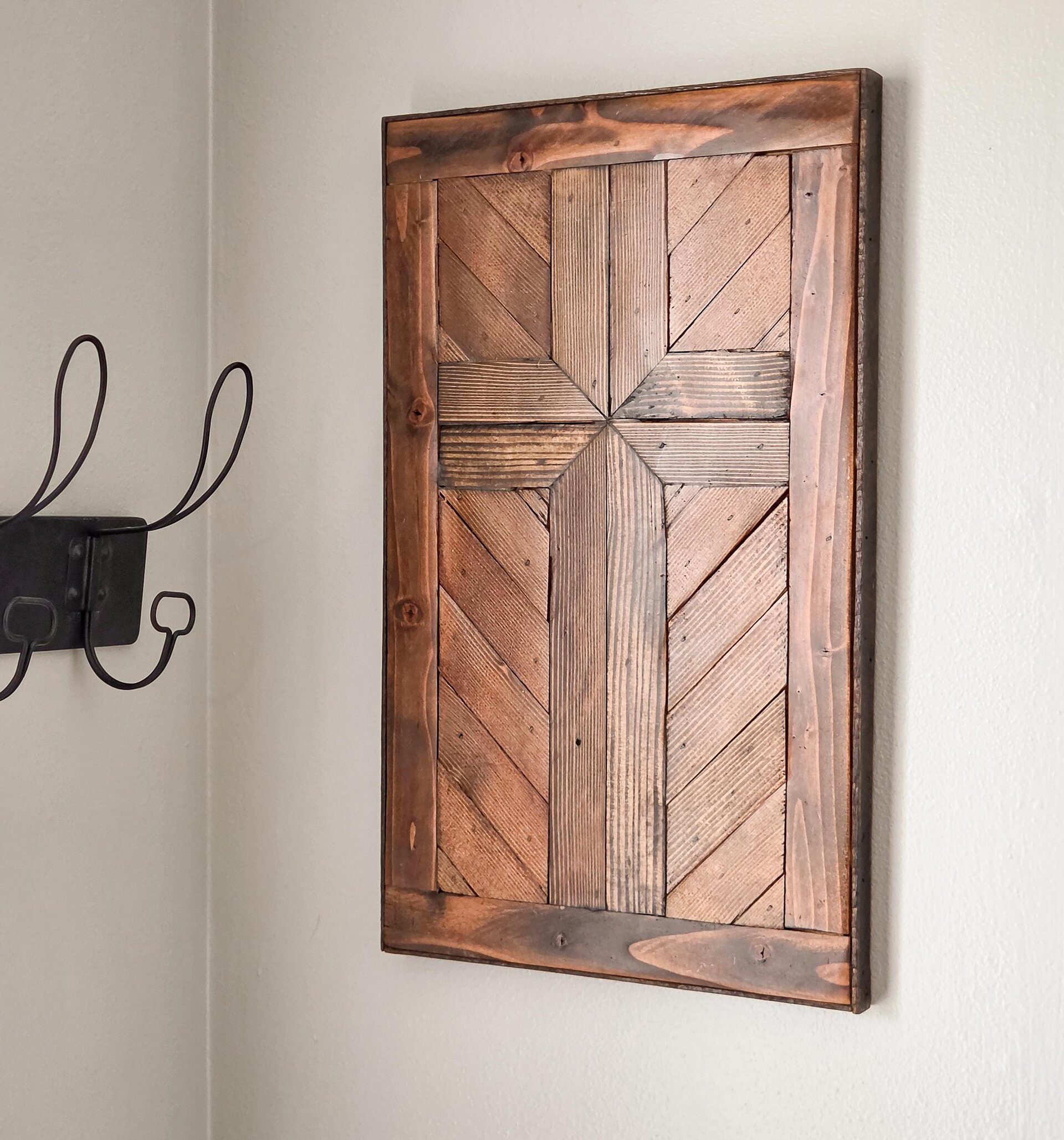 May include: A rectangular wall art piece constructed from dark stained wooden planks arranged in a geometric pattern to form a central cross shape. The wood features visible grain and knots, framed by a solid dark wood border with a smooth finish.