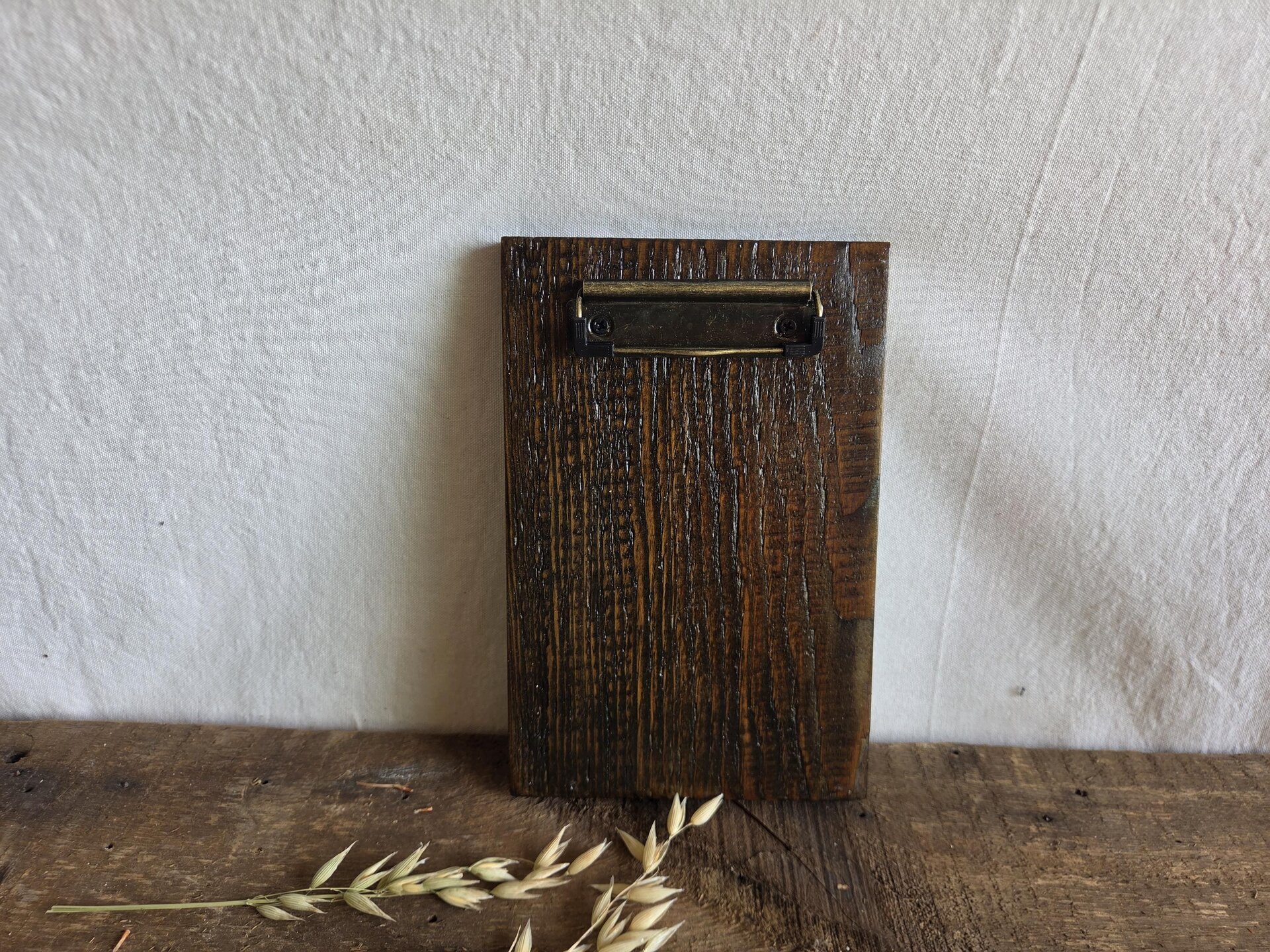 May include: A rectangular clipboard made of dark brown stained wood with a prominent vertical grain texture. The top of the board features a metallic, antique brass-colored spring-loaded clip mechanism secured with two small black screws.