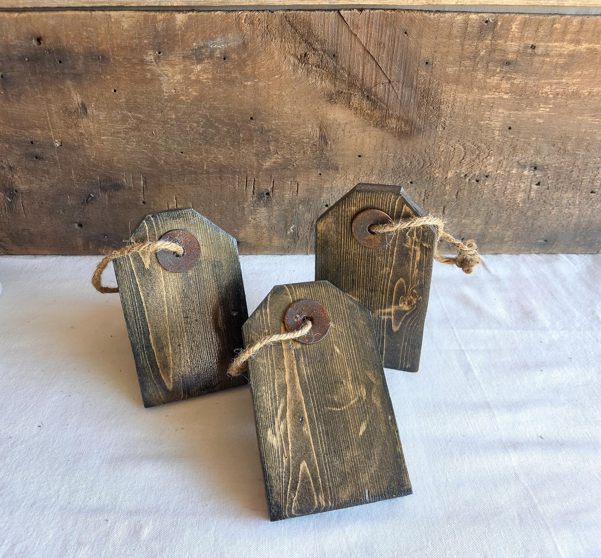 May include: Three rectangular wooden tags with a dark stained finish and visible wood grain. Each tag features a beveled top edge, a circular rusted metal washer centered near the top, and a piece of natural jute twine looped through the hole.