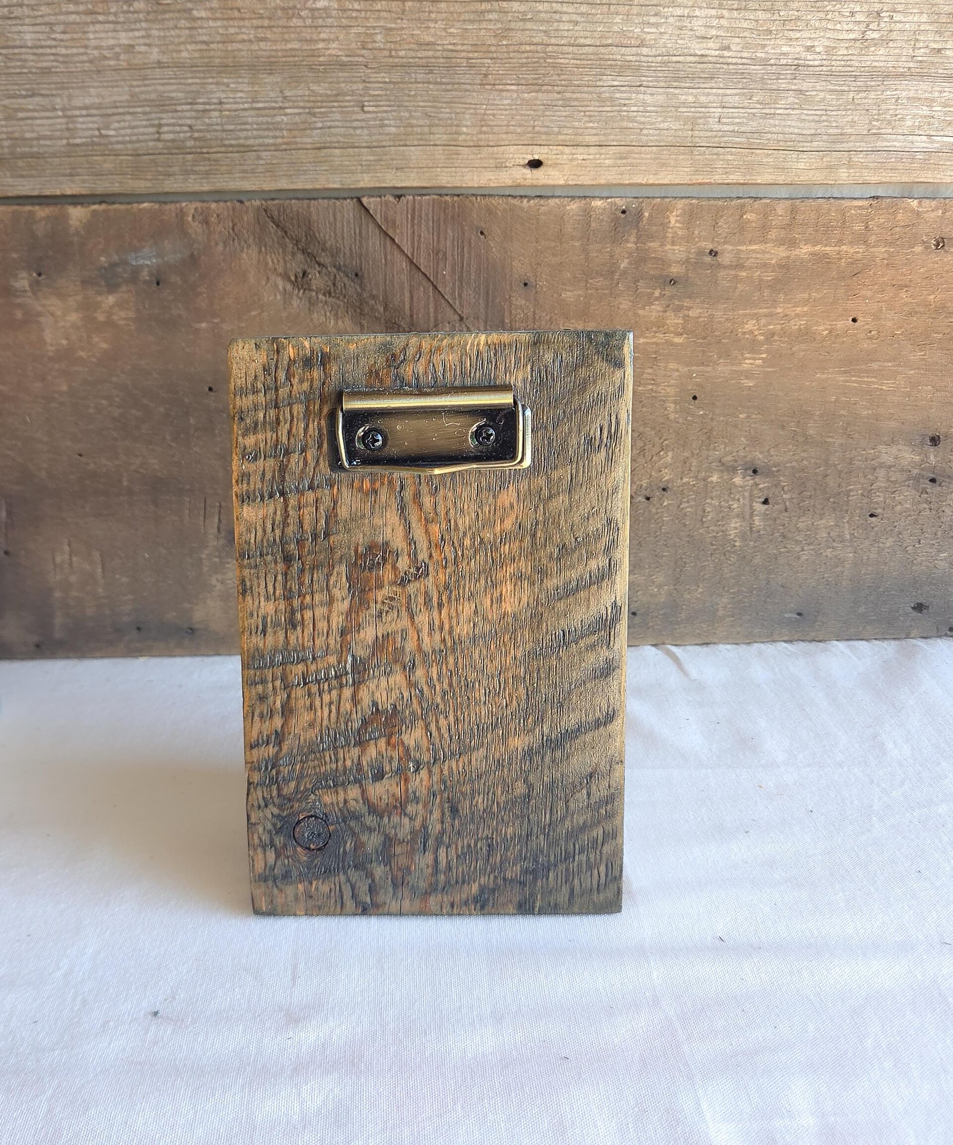 May include: A rectangular piece of reclaimed wood featuring a prominent grain pattern and a dark brown finish. A metallic, brass-toned clip is securely attached to the upper center of the wooden board with two small black screws.