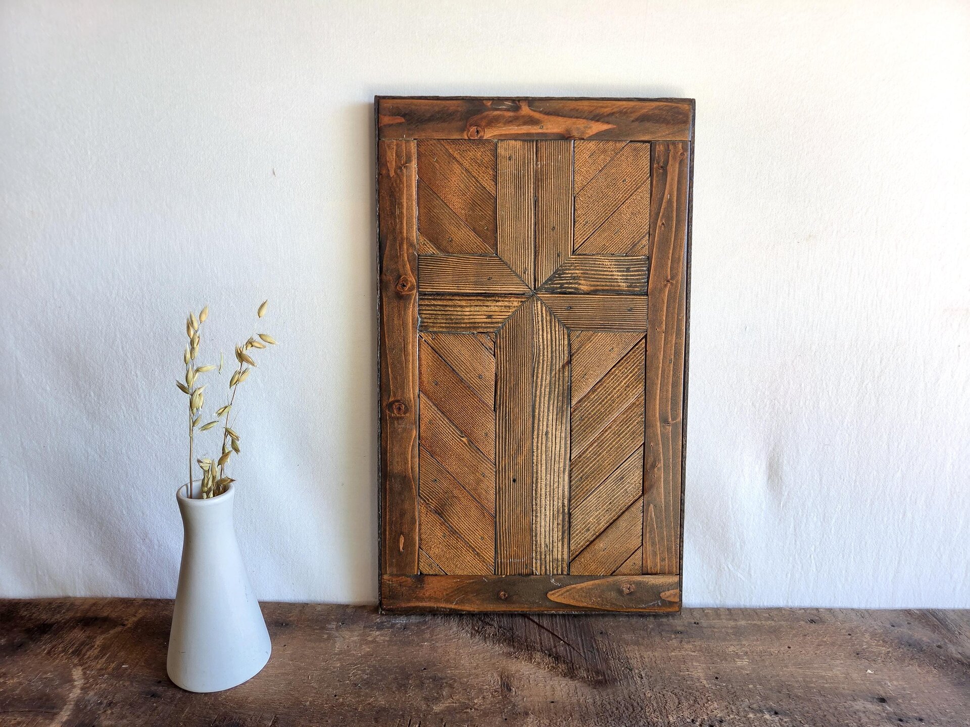 May include: A rectangular wall art piece constructed from dark brown stained wooden planks arranged in a geometric cross pattern. The wood features visible grain and varied textures, framed by a solid border. The design is composed of vertical and diagonal slats.