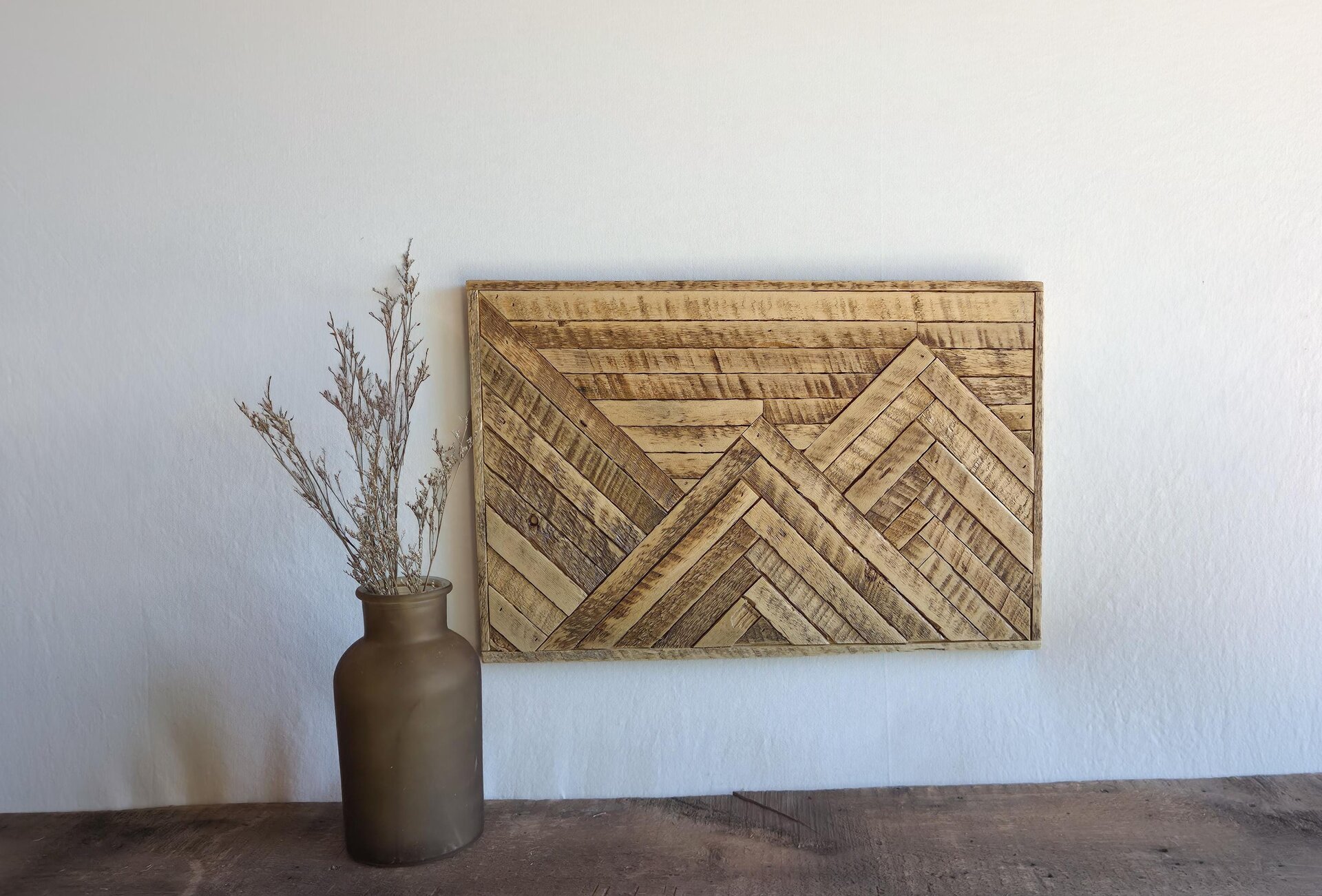 May include: A rectangular wall art piece constructed from light and dark brown wooden slats arranged in a geometric mountain pattern. Beside it sits a matte brown glass vase containing dried, light-colored, branching botanical stems.