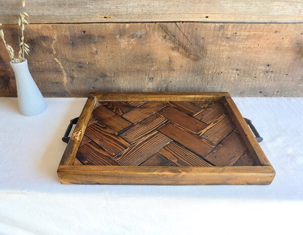 May include: A rectangular wooden serving tray featuring a dark brown herringbone pattern on the base, a thick stained wood frame, and two black metal handles attached to the sides. The tray is constructed from individual wood planks.