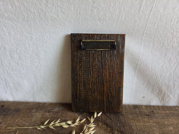 May include: A rectangular clipboard made of dark brown stained wood with a prominent vertical grain texture. The top of the board features a metallic, antique brass-colored spring-loaded clip mechanism secured with two small black screws.