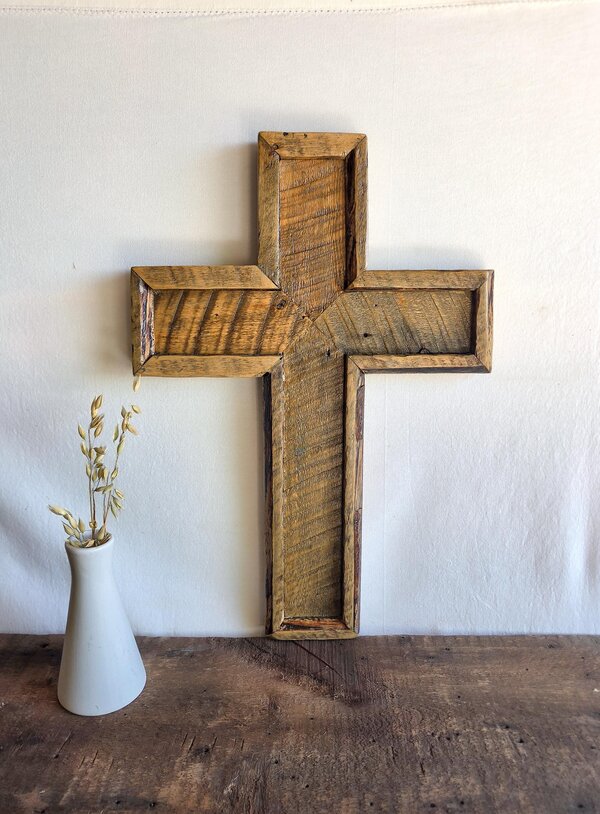 May include: A wall-mounted cross constructed from reclaimed wood with a natural brown finish and visible grain patterns. The cross features a recessed center panel framed by thick wooden borders, showcasing a weathered texture and rustic appearance.
